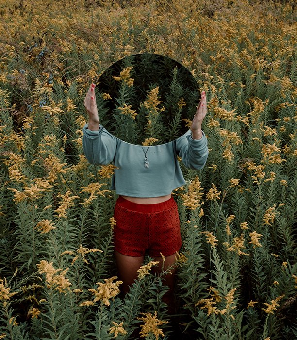 Сюрреалистическая зеркальная фотография женской модели в заросшем поле, держащей зеркало над своим тузом, в котором отражается еще больше заросшей листвы