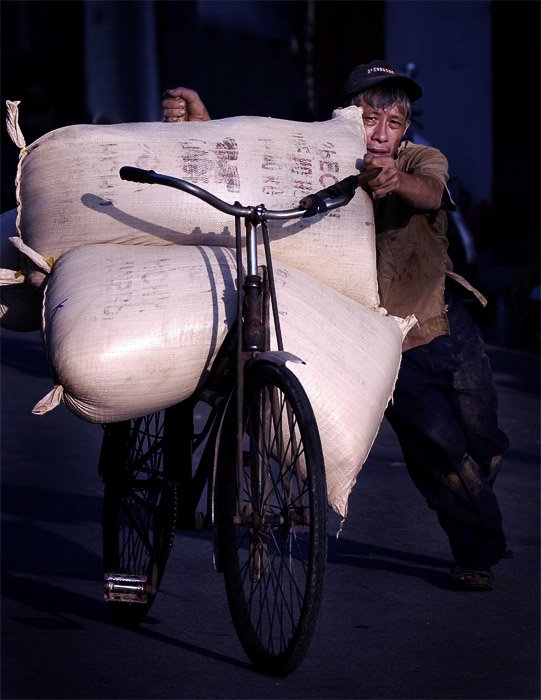 Уличный портрет мужчины, везущего большие мешки риса на велосипеде
