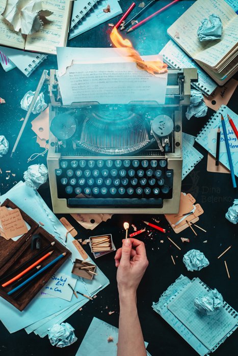 A creative flat lay photo of the messy writing desk and a person holding a match to a typewriter - creative still life photography composition