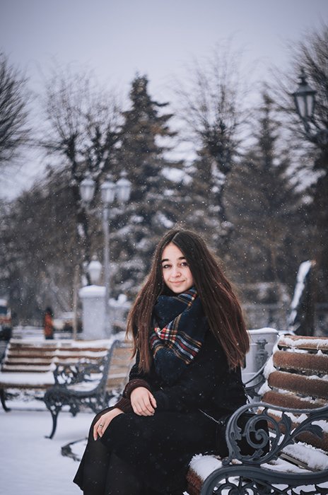 Атмосферный зимний портрет женщины-модели, позирующей на скамейке в падающем снегу