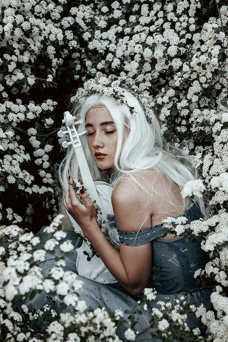 мечтательное портретное изображение беловолосой женщины, приседающей в поле белых цветов и держащей белую скрипку