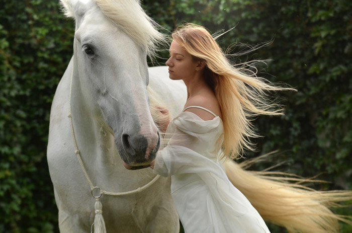 Мечтательная фотография женщины-модели с длинными светлыми волосами, позирующей рядом с белой лошадью