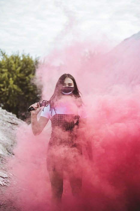 Девушка стоит в розовой дымовой шашке с маской на лице как идея для сюрреалистической фотографии
