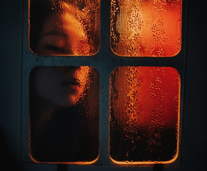 Атмосферный портрет девушки в помещении, прижимающей лицо к оконному стеклу