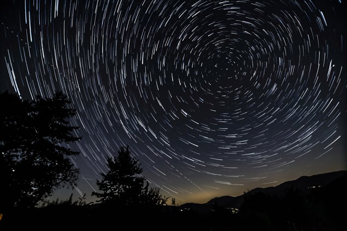 Фотография звездного следа вокруг Северной Полярной звезды с холмами, деревьями и огнями домов.