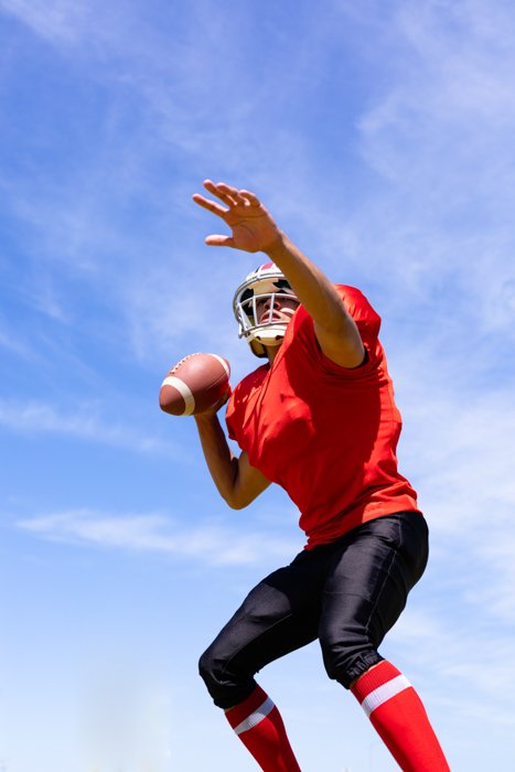 Низкоугольный вид спереди смешанной расы мужчины американского футболиста в форме команды, тренирующегося на спортивном поле, готовящегося бросить футбольный мяч, с голубым небом на заднем плане