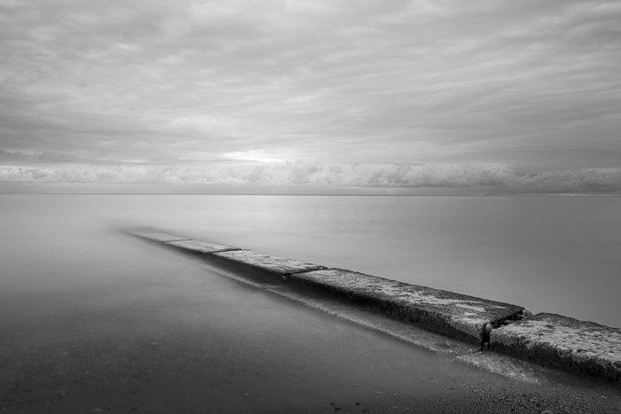 Long exposure photograph of barrier stretching into water at sunrise monochrome