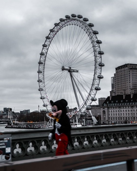 Фотография Лондонского глаза на заднем плане с размытым человеком в костюме Микки Мауса, проходящим мимо на переднем плане