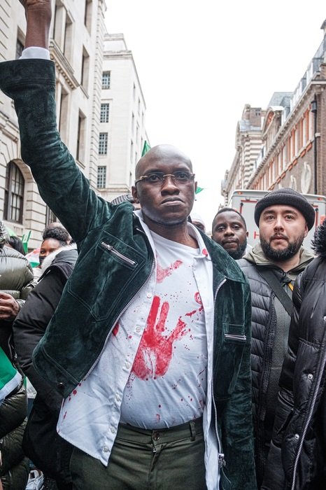 Чернокожий мужчина с поднятой в воздух рукой в знак протеста идет с группой людей по улице